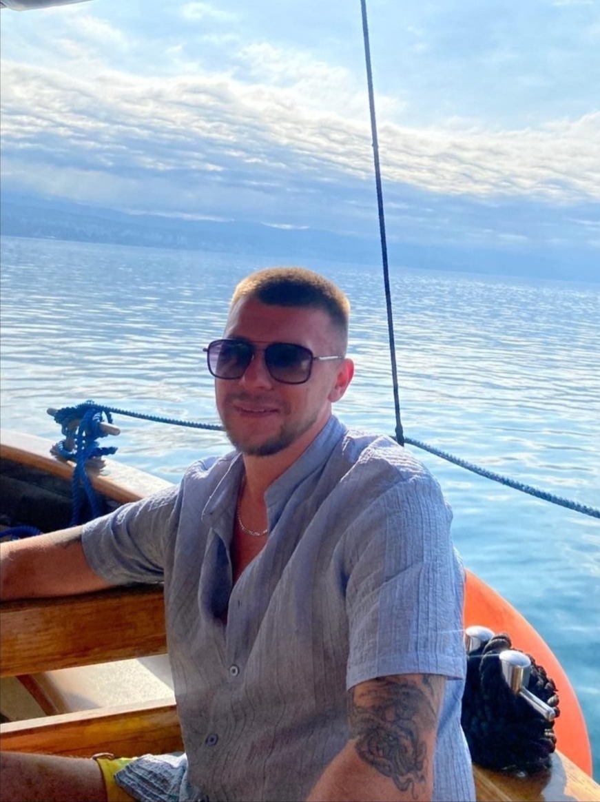 Uśmiechnięty mężczyzna w okularach przeciwsłonecznych, siedzący na łodzi na tle morza i błękitnego nieba z chmurami. Widoczny tatuaż na ramieniu.