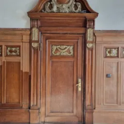 Drewniane drzwi z dekoracyjnymi zdobieniami w stylu retro, otoczone boazerią. Widoczne detale architektoniczne i charakterystyczna kolorystyka drewna.