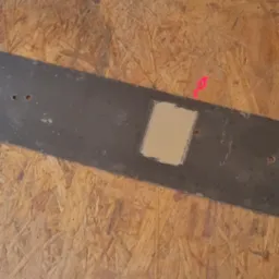 Fragment metalowej listwy z otworami, częściowo oczyszczonej z rdzy, leżącej na płycie OSB. Widoczny ślad po taśmie malarskiej.