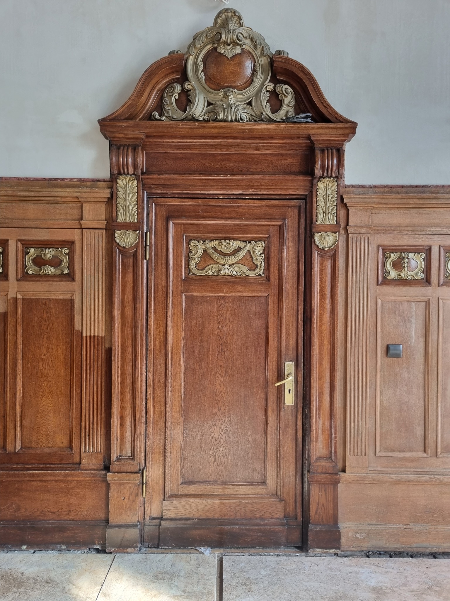 Drewniane drzwi z dekoracyjnymi zdobieniami w stylu retro, otoczone boazerią. Widoczne detale architektoniczne i charakterystyczna kolorystyka drewna.