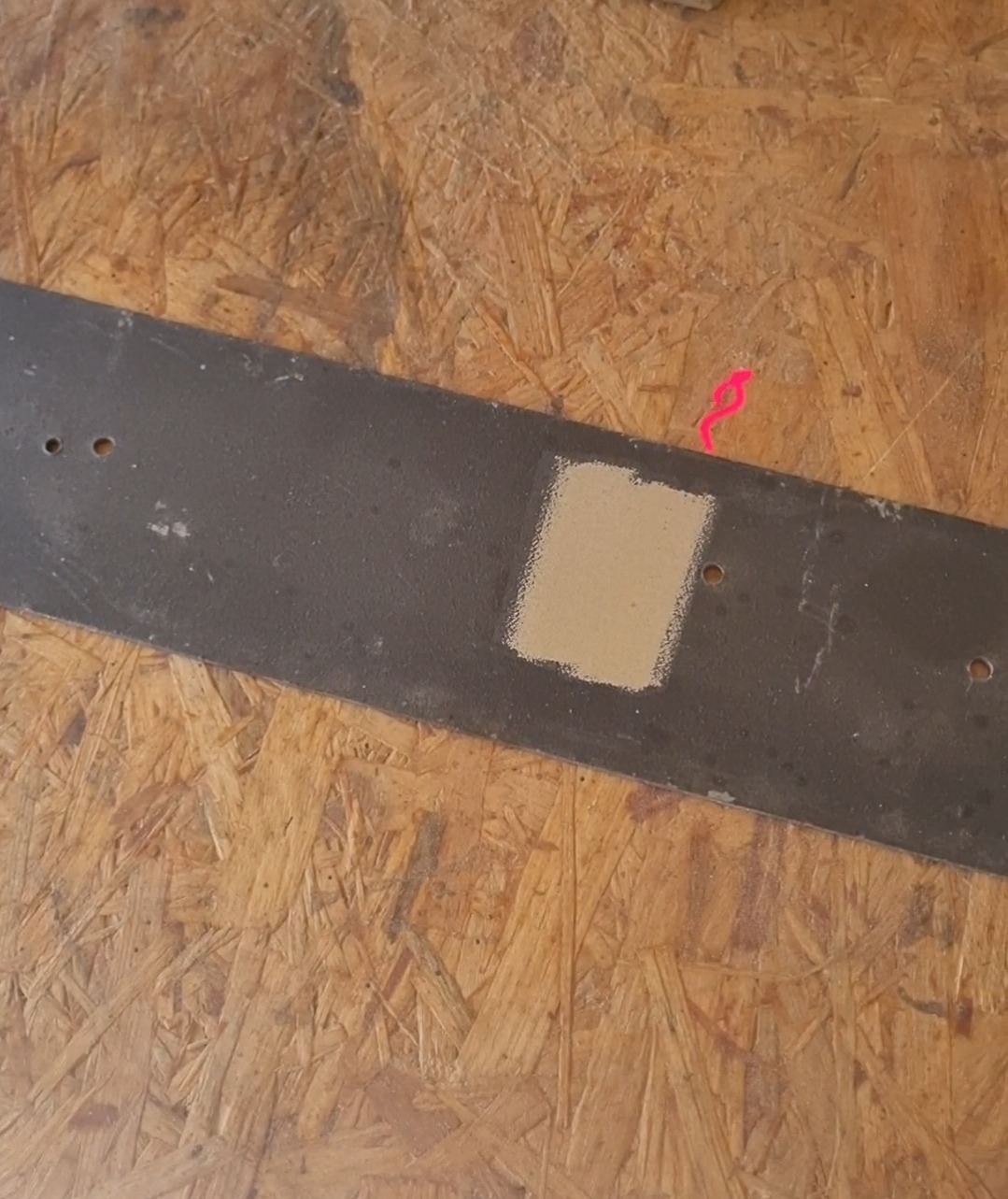 Fragment metalowej listwy z otworami, częściowo oczyszczonej z rdzy, leżącej na płycie OSB. Widoczny ślad po taśmie malarskiej.