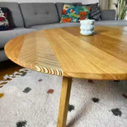 Okrągły, jasny stolik kawowy z drewna dębowego z unikalnym wzorem na krawędzi blatu, ustawiony na dywanie w salonie. W tle szara sofa i dekoracyjna ceramika.