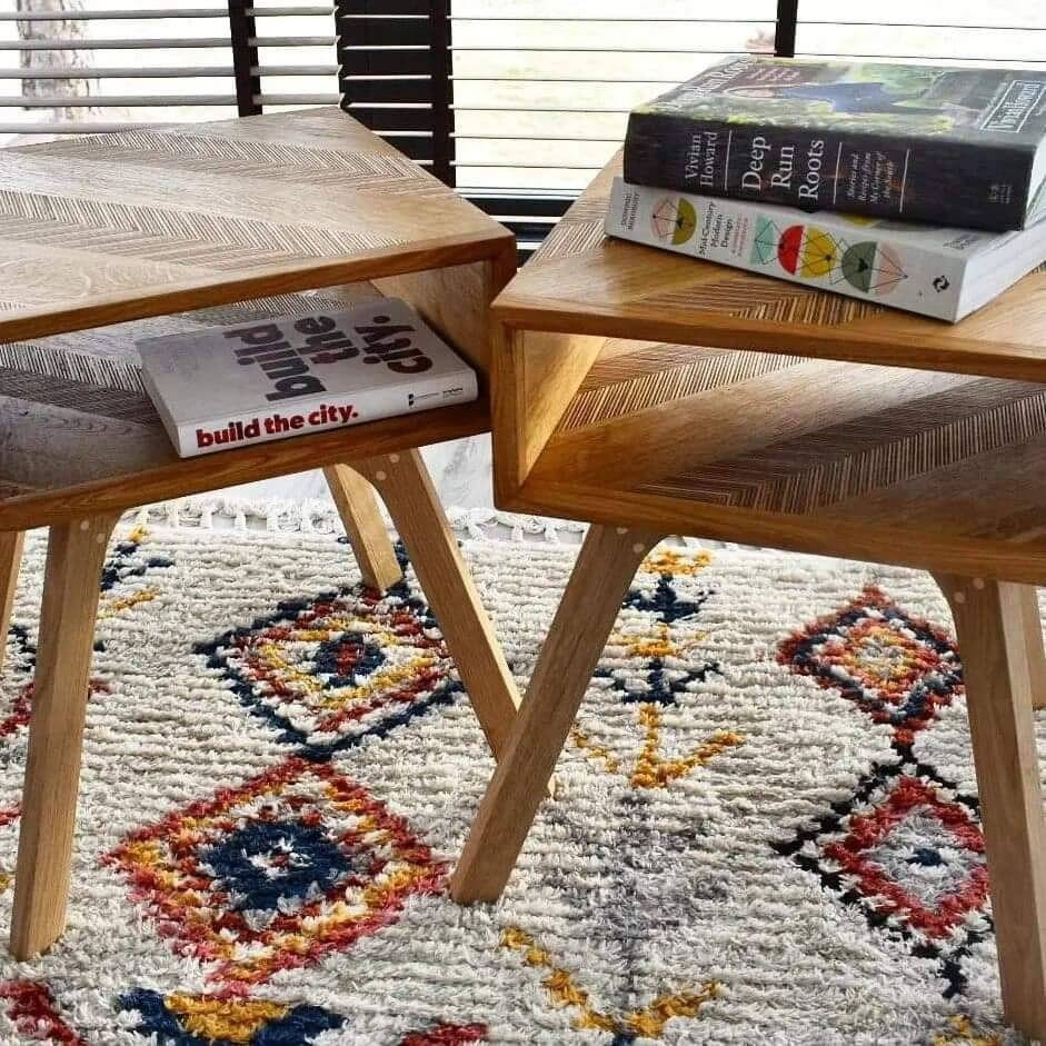 Para drewnianych stolików nocnych z półkami, ustawionych na wzorzystym dywanie. Blaty stolików zdobi subtelny wzór jodełki. Na blatach leżą książki.