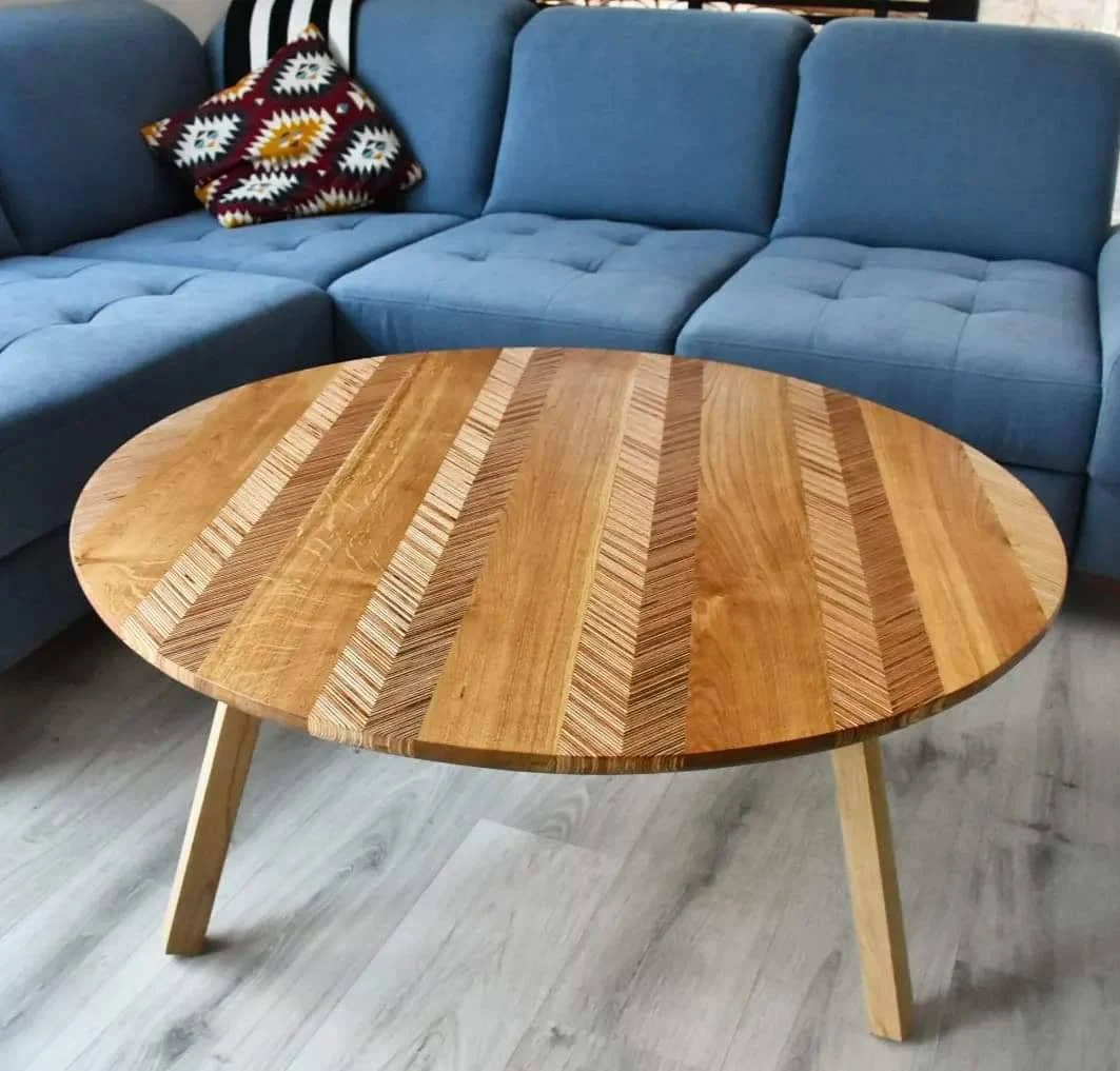 Okrągły stolik kawowy z blatem z drewna w jodełkę, na tle niebieskiej sofy. Jasne drewniane nogi i parkiet. Unikalny wzór słojów.