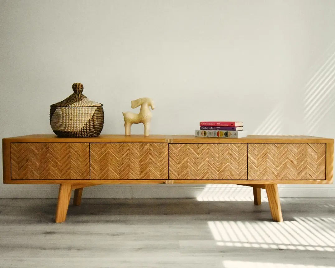 Drewniana szafka RTV z czterema frontami z wzorem w jodełkę. Na blacie koszyk, figurka i książki. Całość oświetlona promieniami słońca padającymi przez żaluzje.