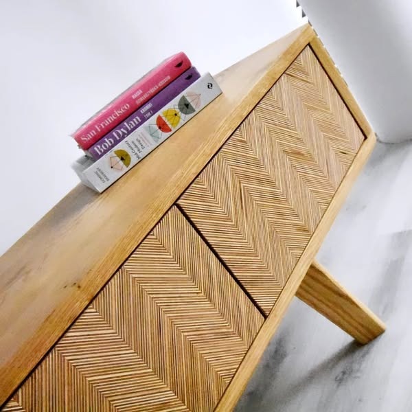 Drewniana komoda z wzorem jodełki na frontach szuflad, ustawiona pod kątem, z książkami na blacie. Naturalne oświetlenie podkreśla strukturę drewna.