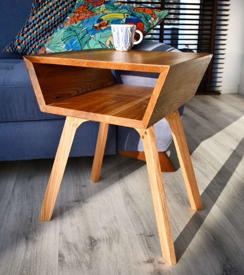 Drewniany stolik kawowy o modernistycznym designie, z półką i skośnymi krawędziami, ustawiony na tle sofy z wzorzystymi poduszkami. Jasne drewno, naturalne światło.