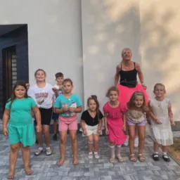 Grupa dzieci z animatorką w różowej tiulowej spódnicy pozuje na tle nowoczesnego budynku z elementami drewnianymi i kopułą cerkwi w tle.