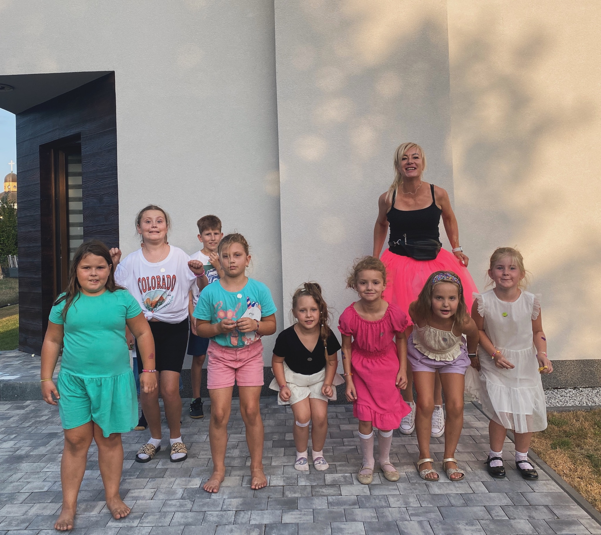 Grupa dzieci z animatorką w różowej tiulowej spódnicy pozuje na tle nowoczesnego budynku z elementami drewnianymi i kopułą cerkwi w tle.