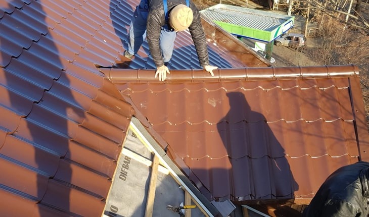 Montaż brązowej blachodachówki modułowej na dachu domu. Widoczny dekarz w trakcie pracy, elementy więźby i membrana dachowa. Ujęcie z góry, w tle budynek stacji benzynowej.