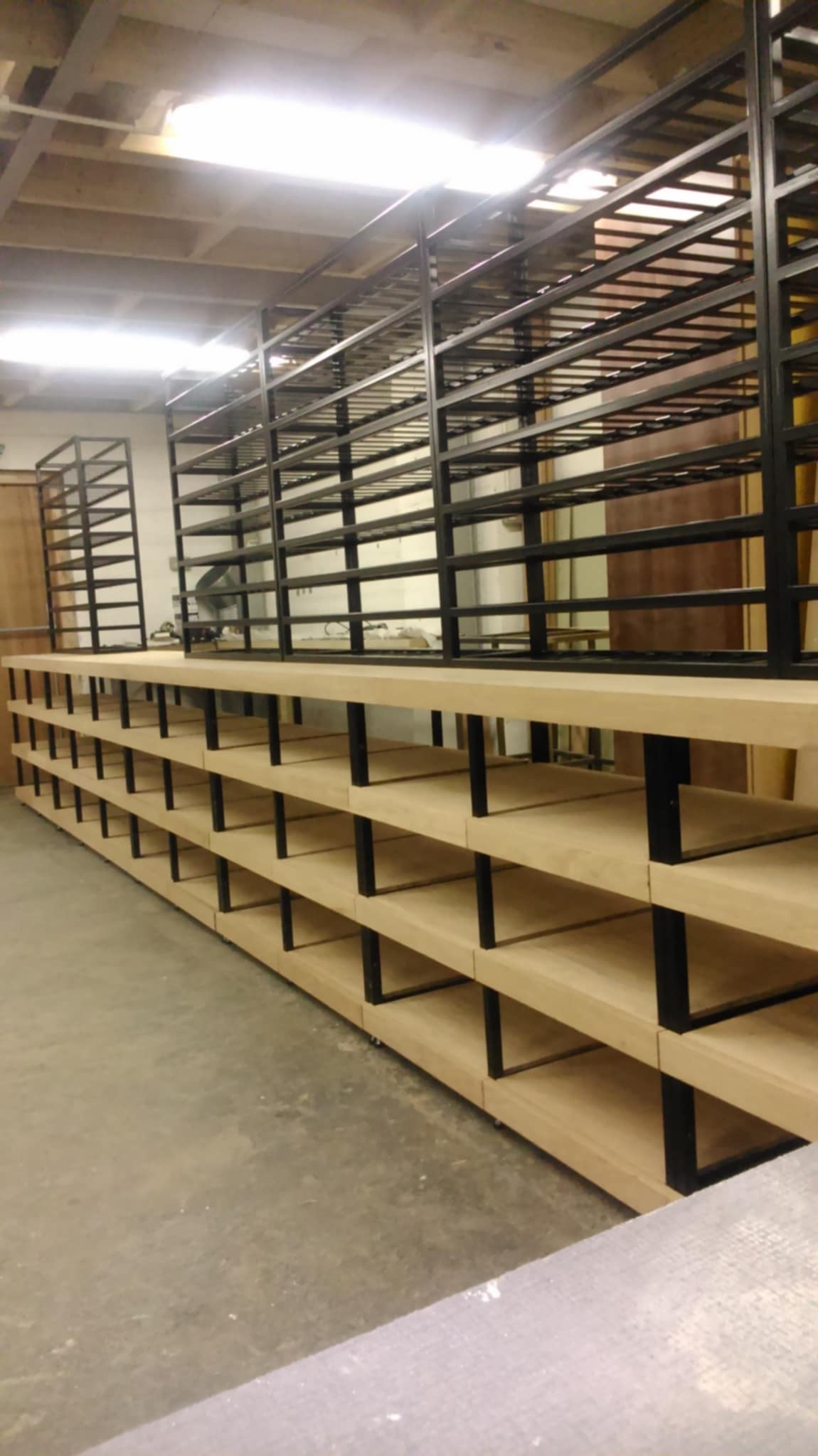 Metalowa konstrukcja regału z drewnianymi półkami w warsztacie, widok z perspektywy. Czarna rama, jasne drewno, surowe oświetlenie.