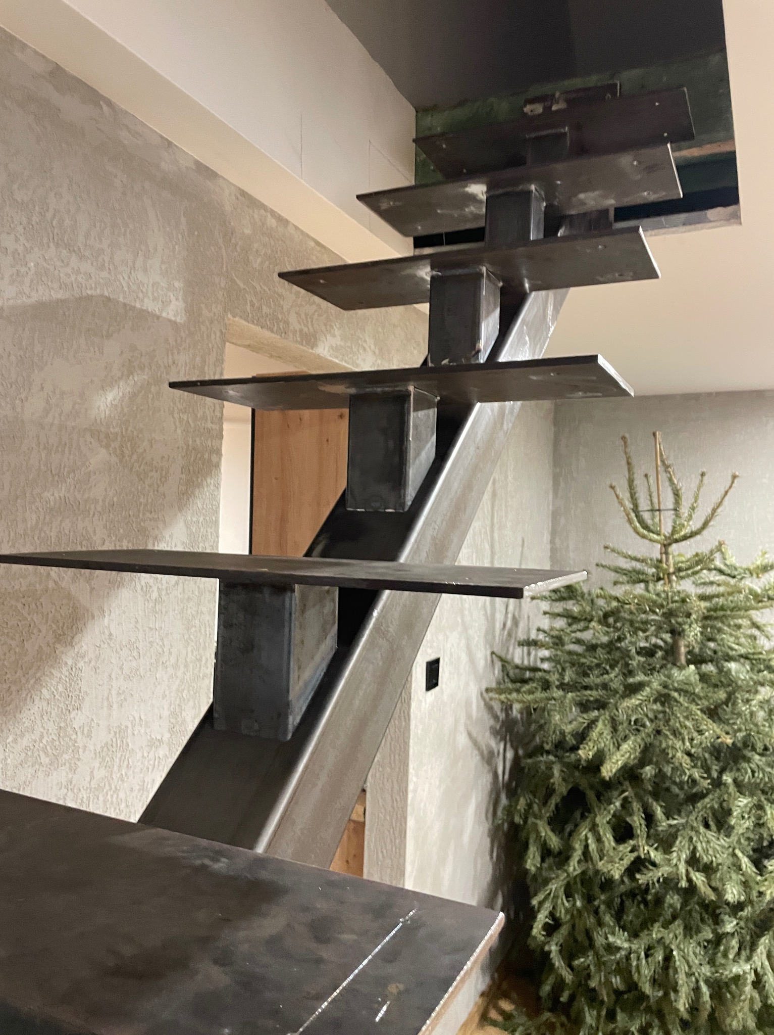 Surowe, metalowe schody ażurowe z kwadratowymi stopniami, widoczne z dołu, z choinką w tle. Minimalistyczny design.