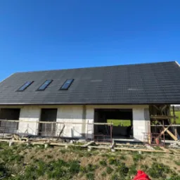 Dom w stanie surowym, z czarnym dachem z dachówki i trzema oknami dachowymi, na tle błękitnego nieba i zielonego krajobrazu. Widoczne rusztowania i materiały budowlane.