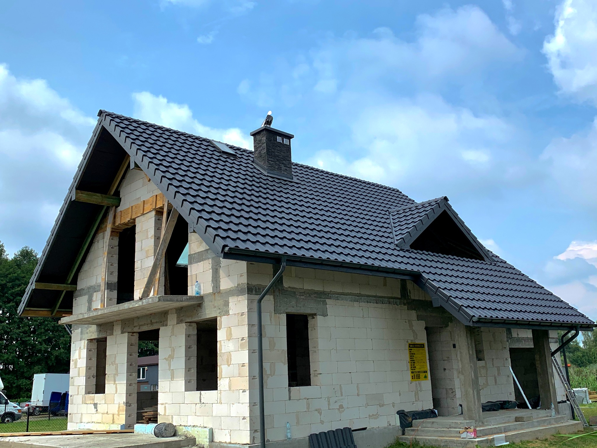 Dom w trakcie budowy z czarnym dachem, na kominie siedzi figurka orła. Widoczne pustaki i elementy konstrukcji dachu na tle błękitnego nieba.