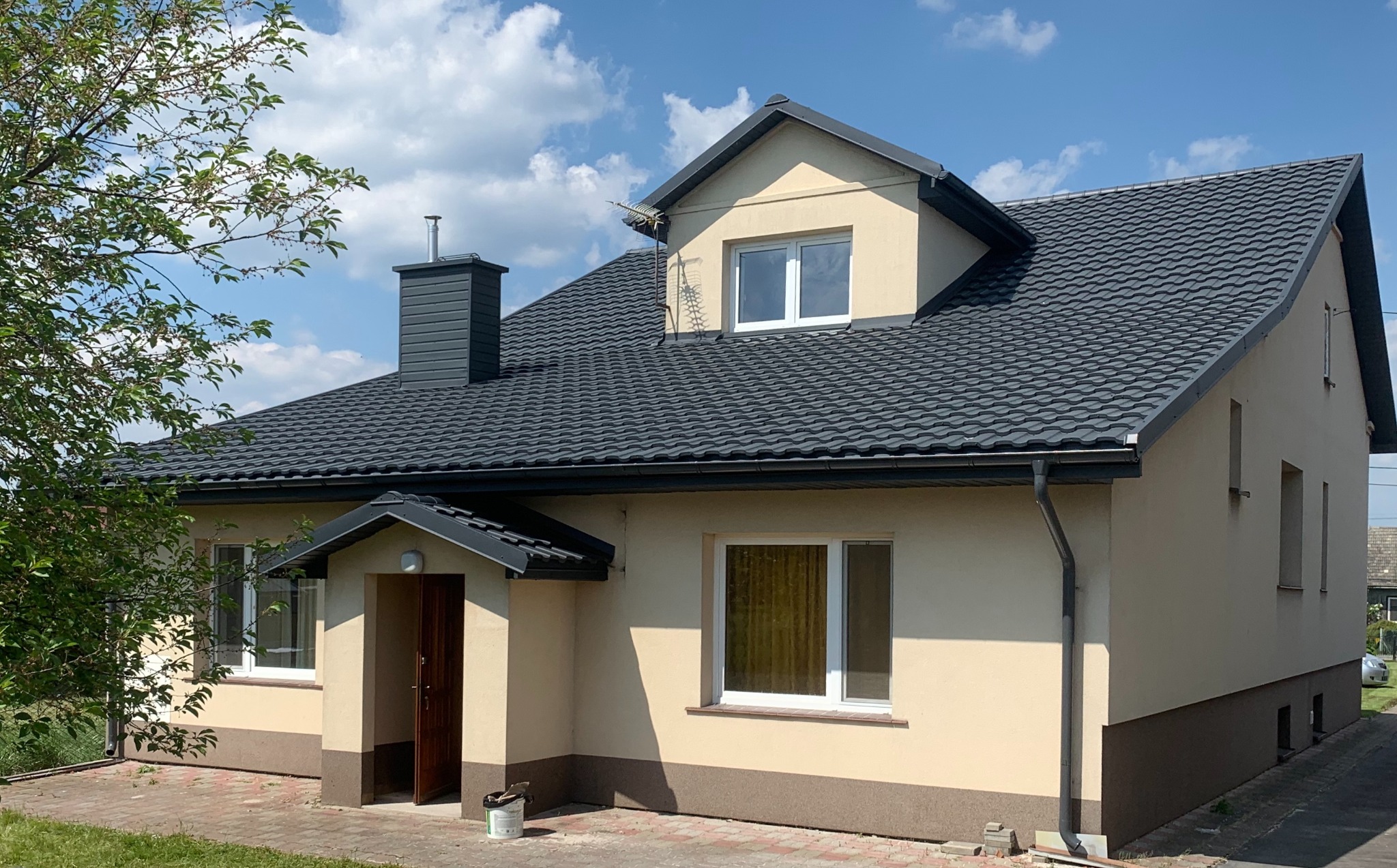 Dom jednorodzinny z nowym, grafitowym dachem z blachodachówki, ciemnymi obróbkami i kominem. Na pierwszym planie drzewo i wiadro po farbie.