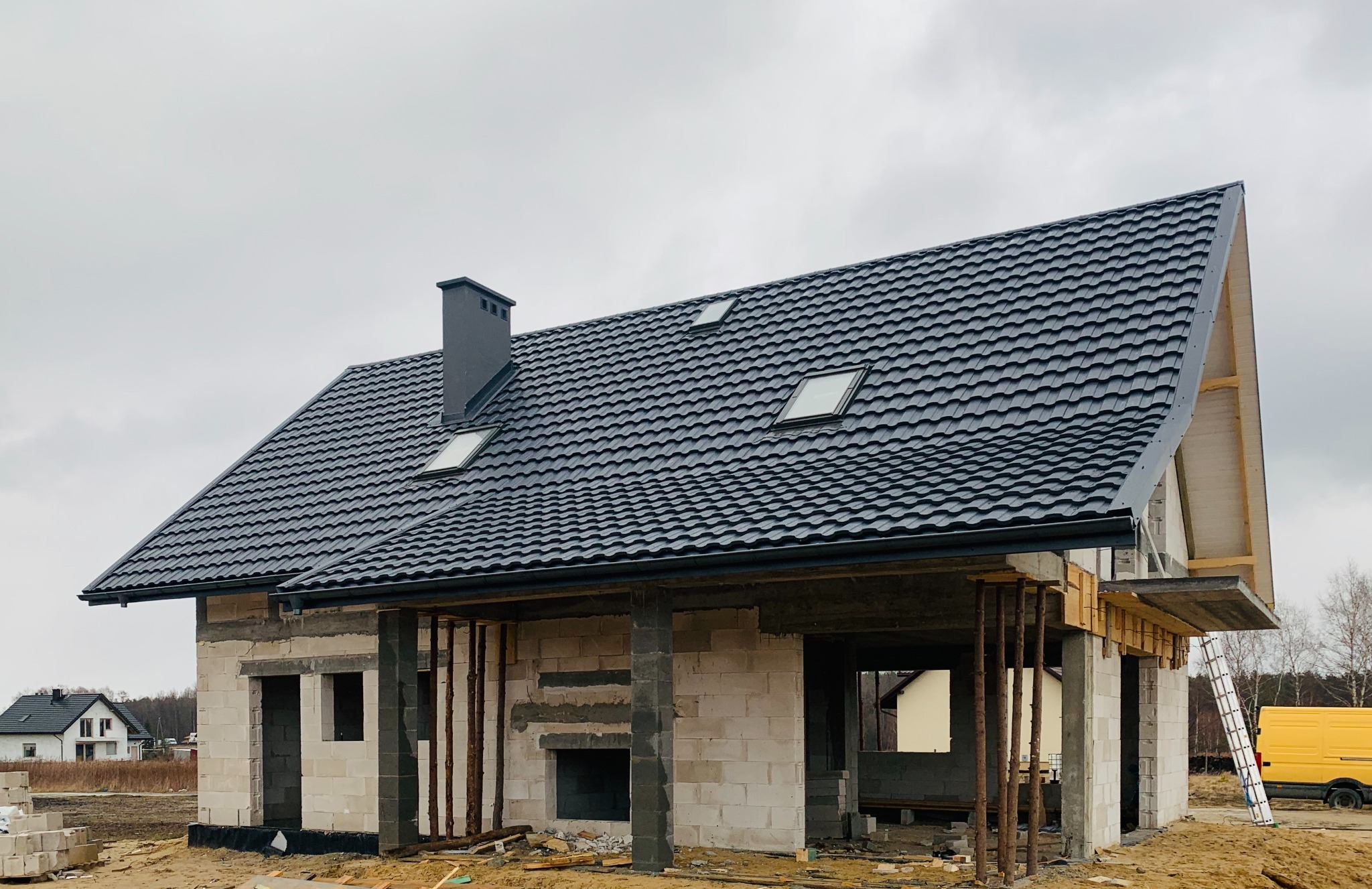 Dom w trakcie budowy, z czarnym dachem z blachodachówki i szarym kominem. Widoczne rusztowania i pustaki. Na dachu okna dachowe.