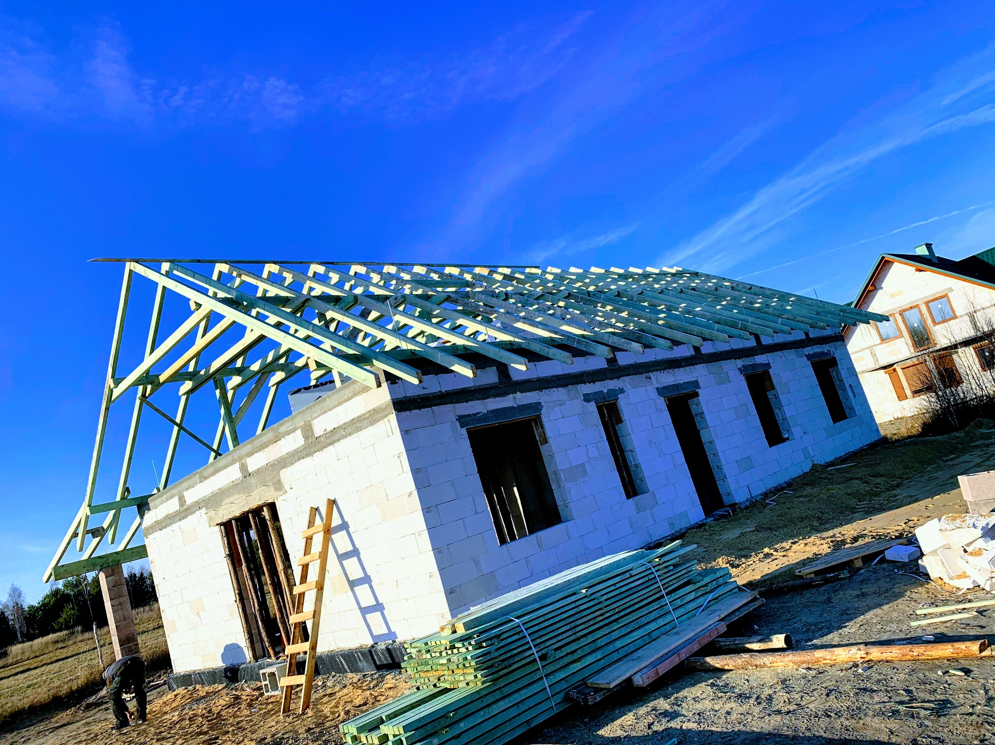 Wznoszenie domu z bloczków: surowe ściany, drewniana więźba dachowa w trakcie montażu, błękitne niebo w tle. Widoczne deski i drabina.