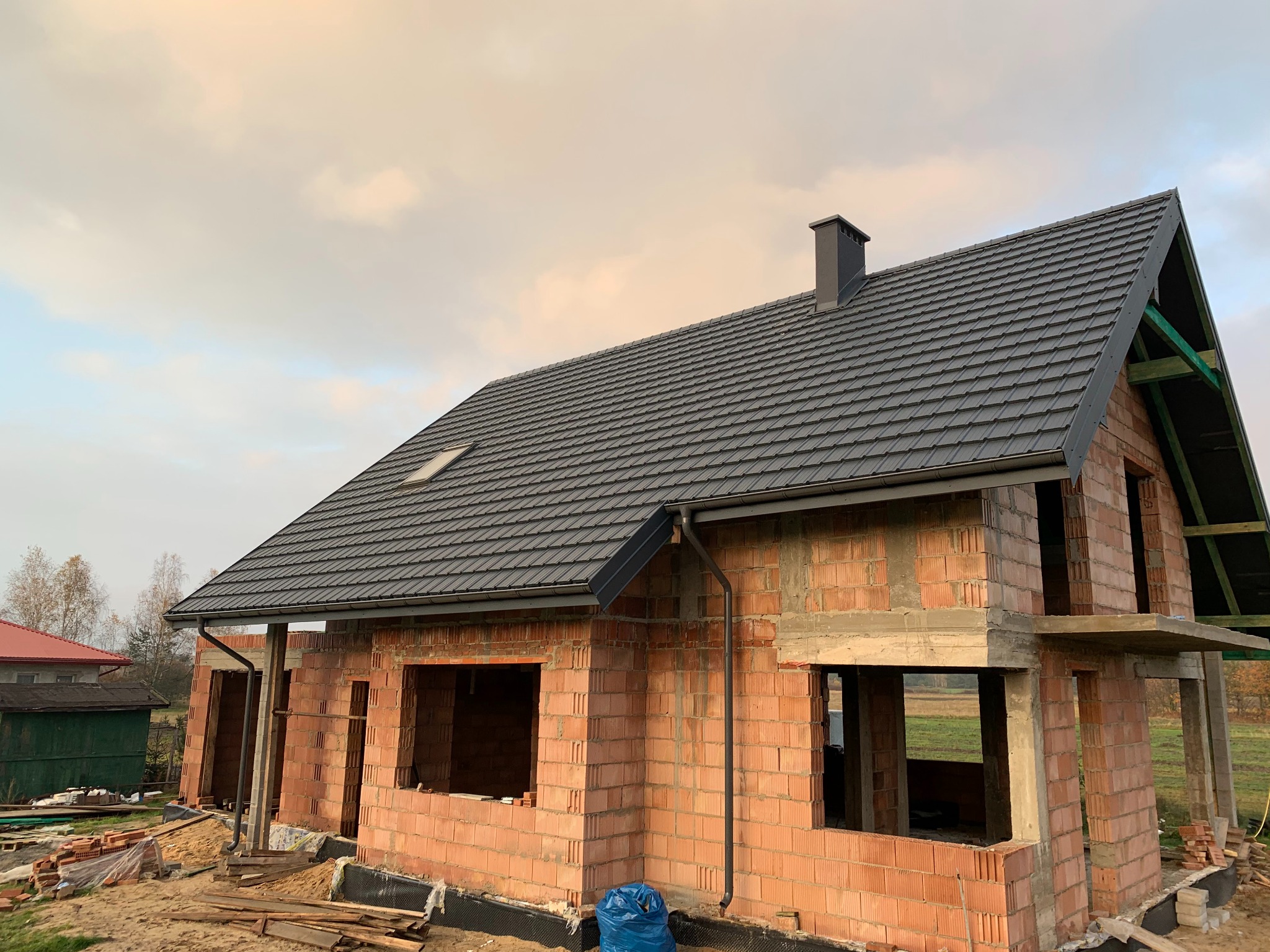Dom w trakcie budowy z ciemnoszarą dachówką, widoczne mury z cegły i elementy konstrukcji dachu. Ujęcie z dołu, pochmurne niebo w tle.