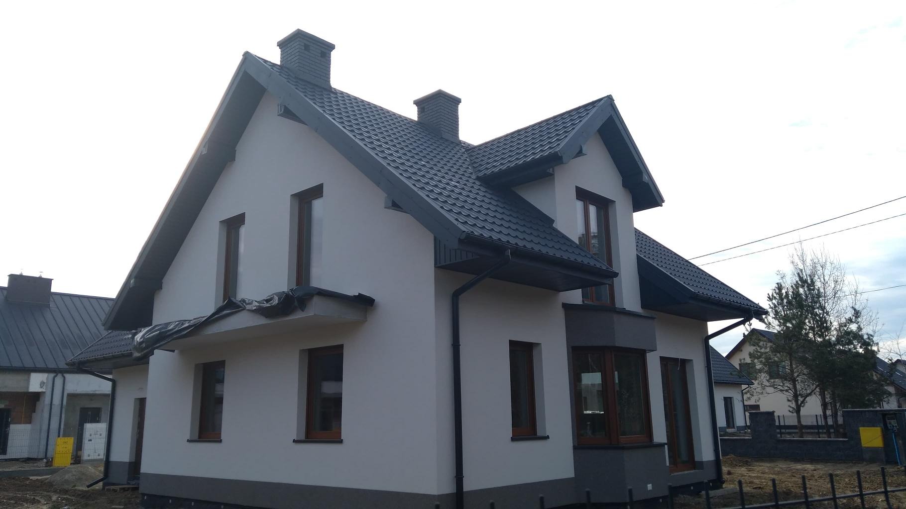 Nowoczesny dom jednorodzinny z ciemną dachówką i szarymi obróbkami blacharskimi, widoczny z zewnątrz, z fragmentem dachu sąsiedniego budynku.