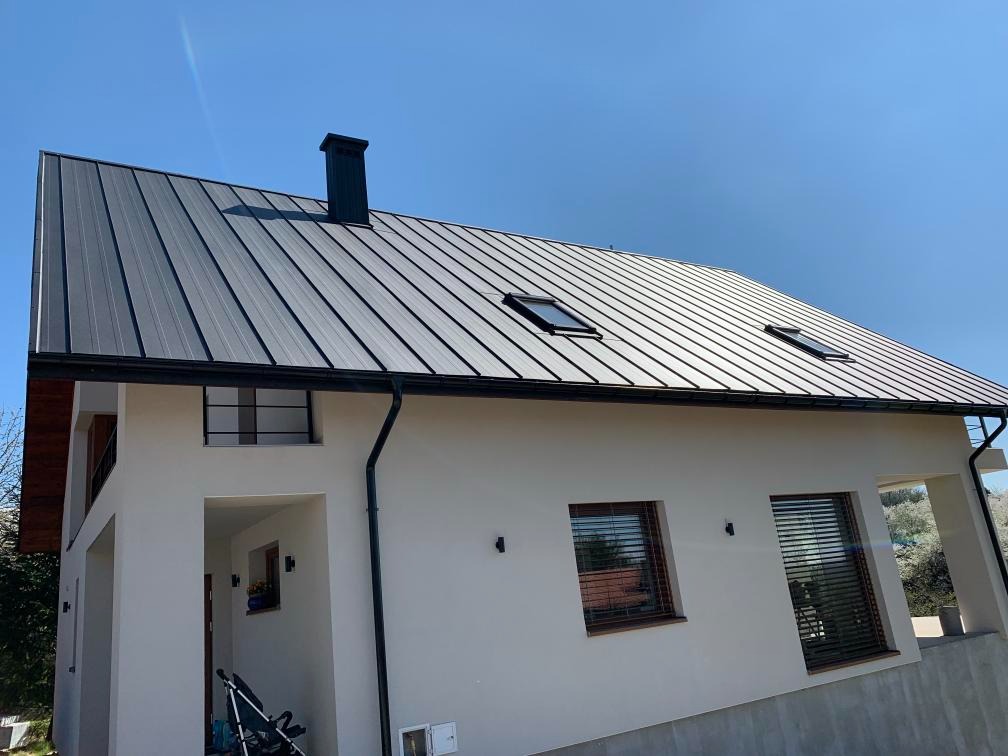 Nowoczesny dom z grafitowym dachem z blachy trapezowej, z oknami dachowymi i czarną obróbką blacharską, na tle błękitnego nieba.