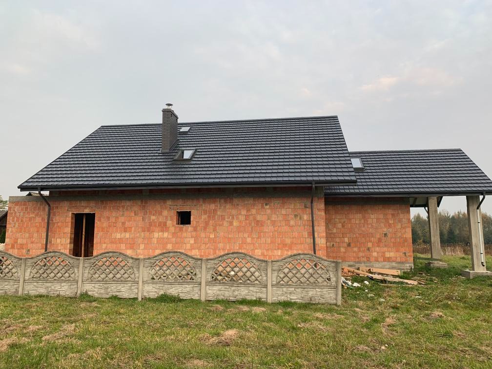 Dom w trakcie budowy z nowym, grafitowym dachem z dachówką, widoczne okna dachowe i komin, ogrodzenie z przodu. Surowy stan budynku.