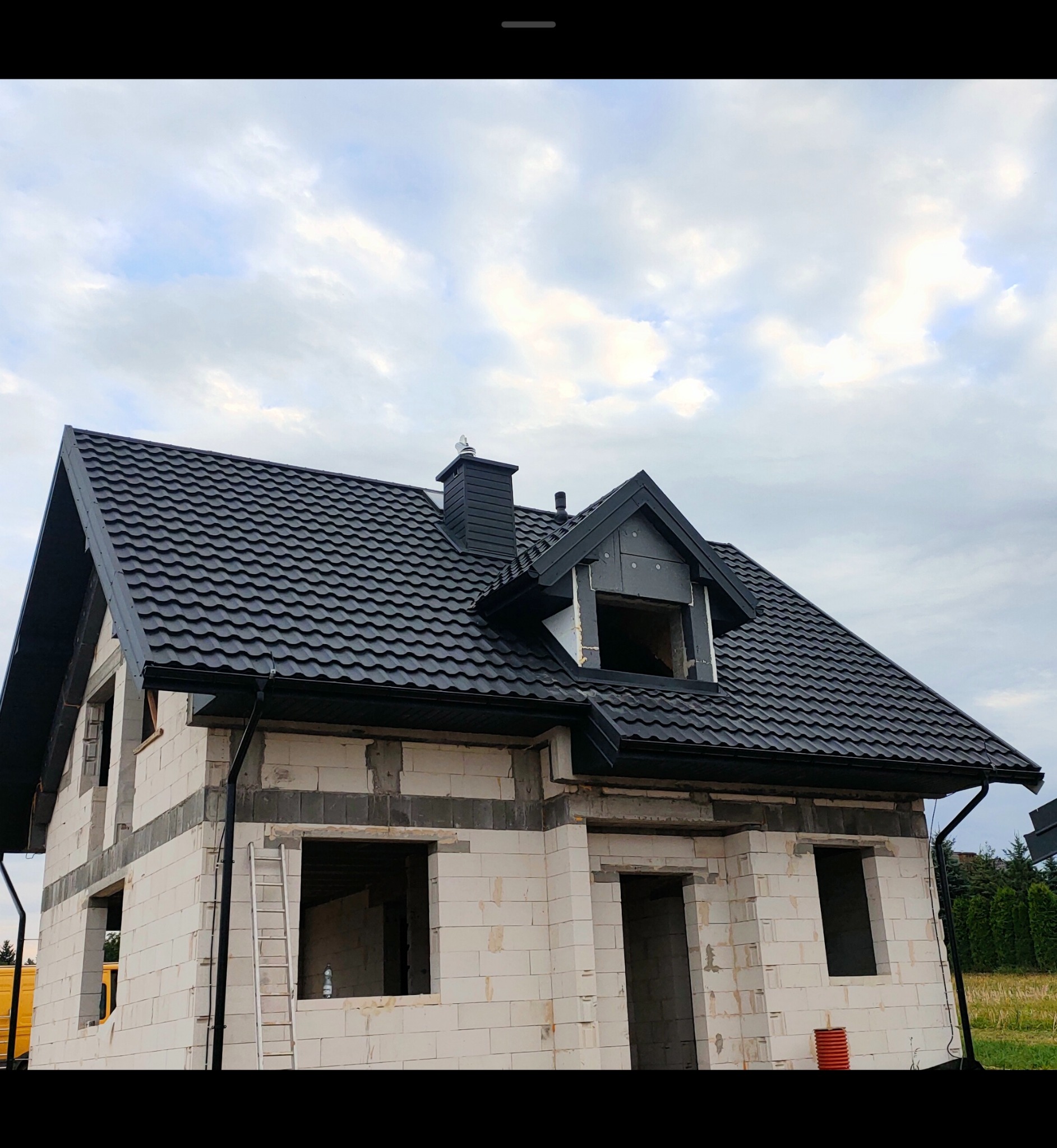 Dom w stanie surowym z czarnym, nowym dachem z blachodachówki. Widoczne orynnowanie i komin. Budynek z pustaków pod błękitnym niebem.