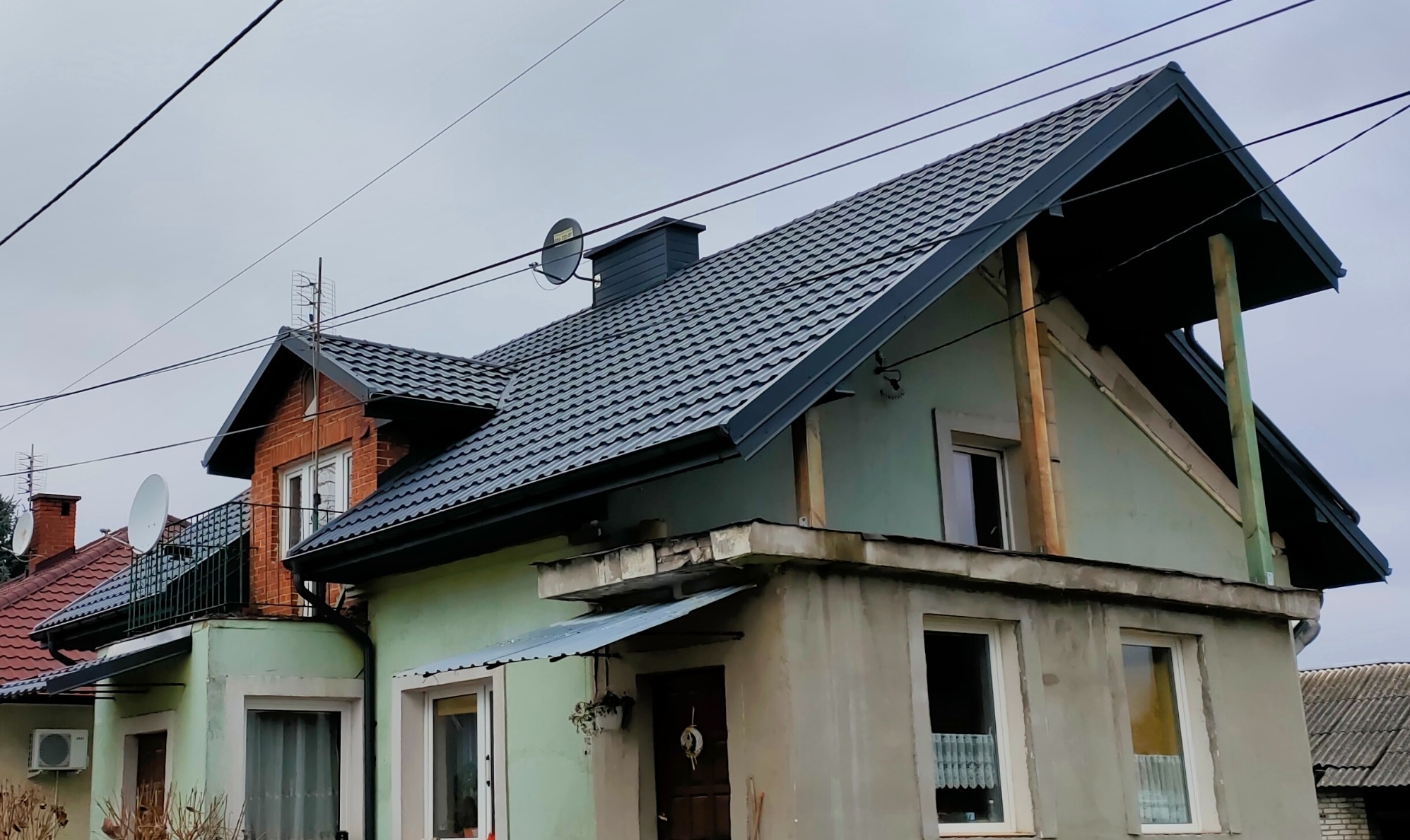 Dom z nowym, grafitowym dachem z blachy trapezowej. Widoczne anteny satelitarne i komin. Część elewacji w trakcie wykończenia.