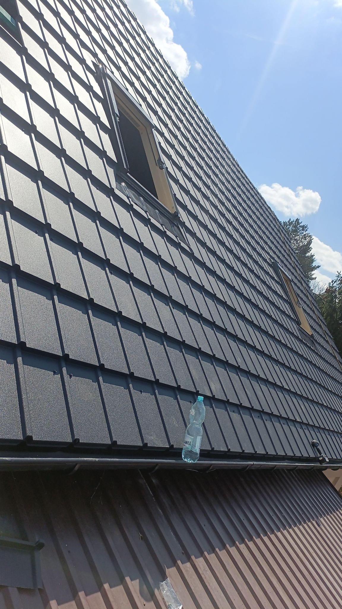 Nowy, grafitowy dach z blachodachówki z dwoma oknami połaciowymi, widziany pod kątem. Na rynnie stoi butelka wody. Błękitne niebo w tle.