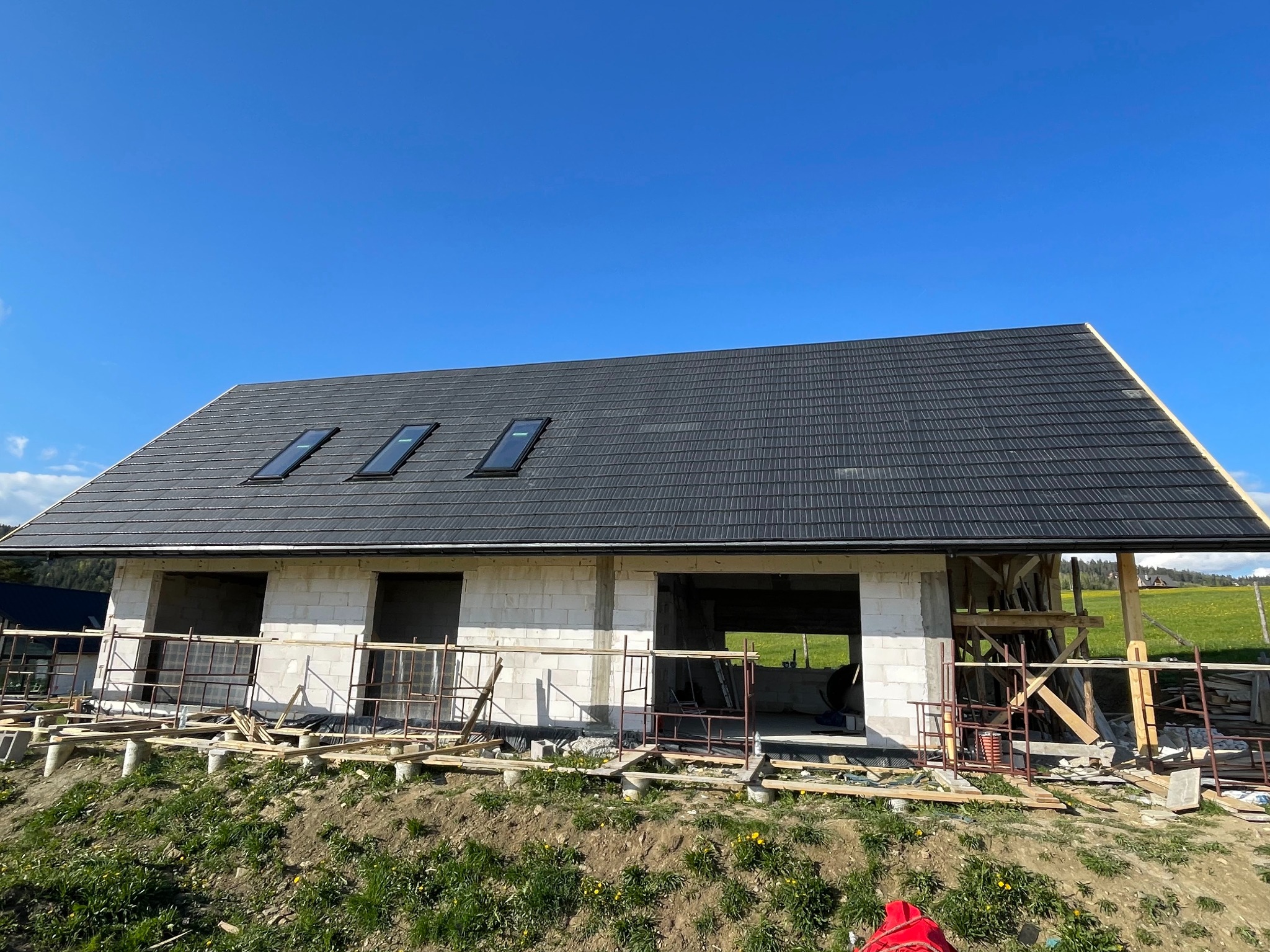 Dom w stanie surowym, z czarnym dachem z dachówki i trzema oknami dachowymi, na tle błękitnego nieba i zielonego krajobrazu. Widoczne rusztowania i materiały budowlane.