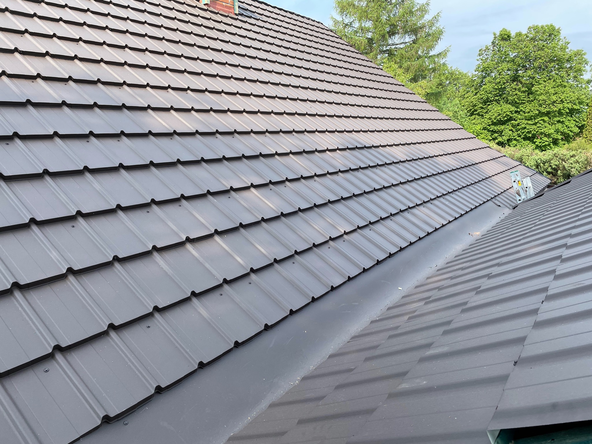 Szary dach z blachy trapezowej, widoczny z boku pod kątem, na tle zieleni drzew i fragmentu nieba. Widoczne łączenia blachy.