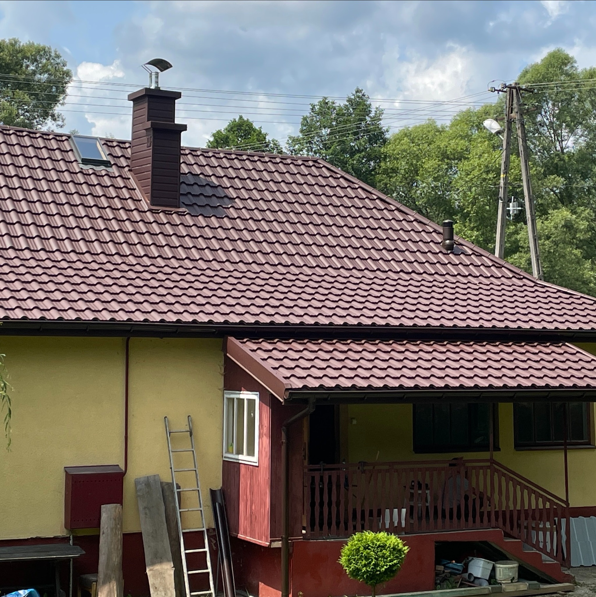 Dom z nowym, bordowym dachem z blachodachówki, brązowym kominem z daszkiem i oknem dachowym. Przy ścianie drabina i elementy drewniane.