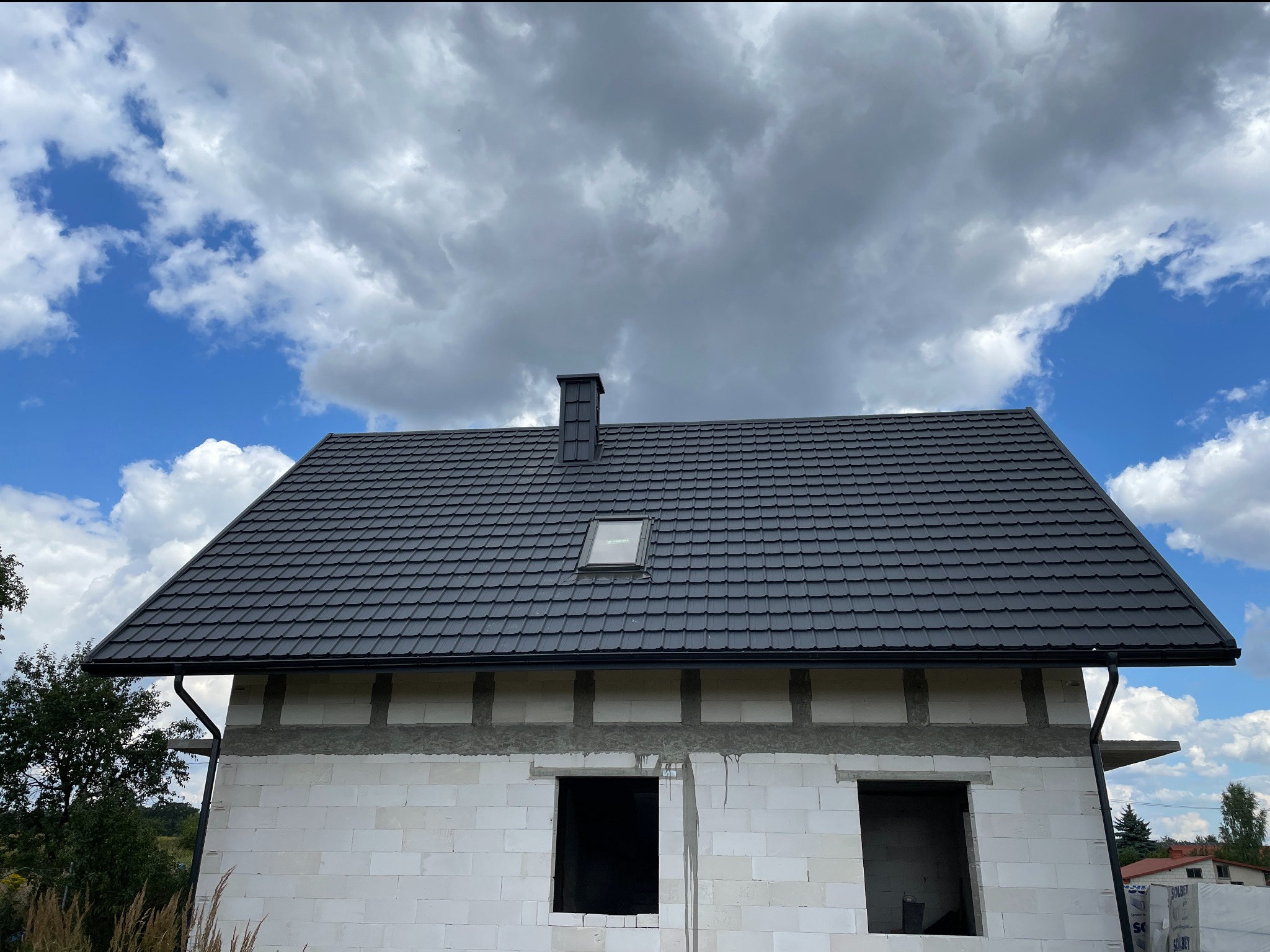Dom w trakcie budowy z ciemnym, nowym dachem z blachy, widoczne okno dachowe i orynnowanie. Białe ściany z betonu komórkowego, błękitne niebo w tle.