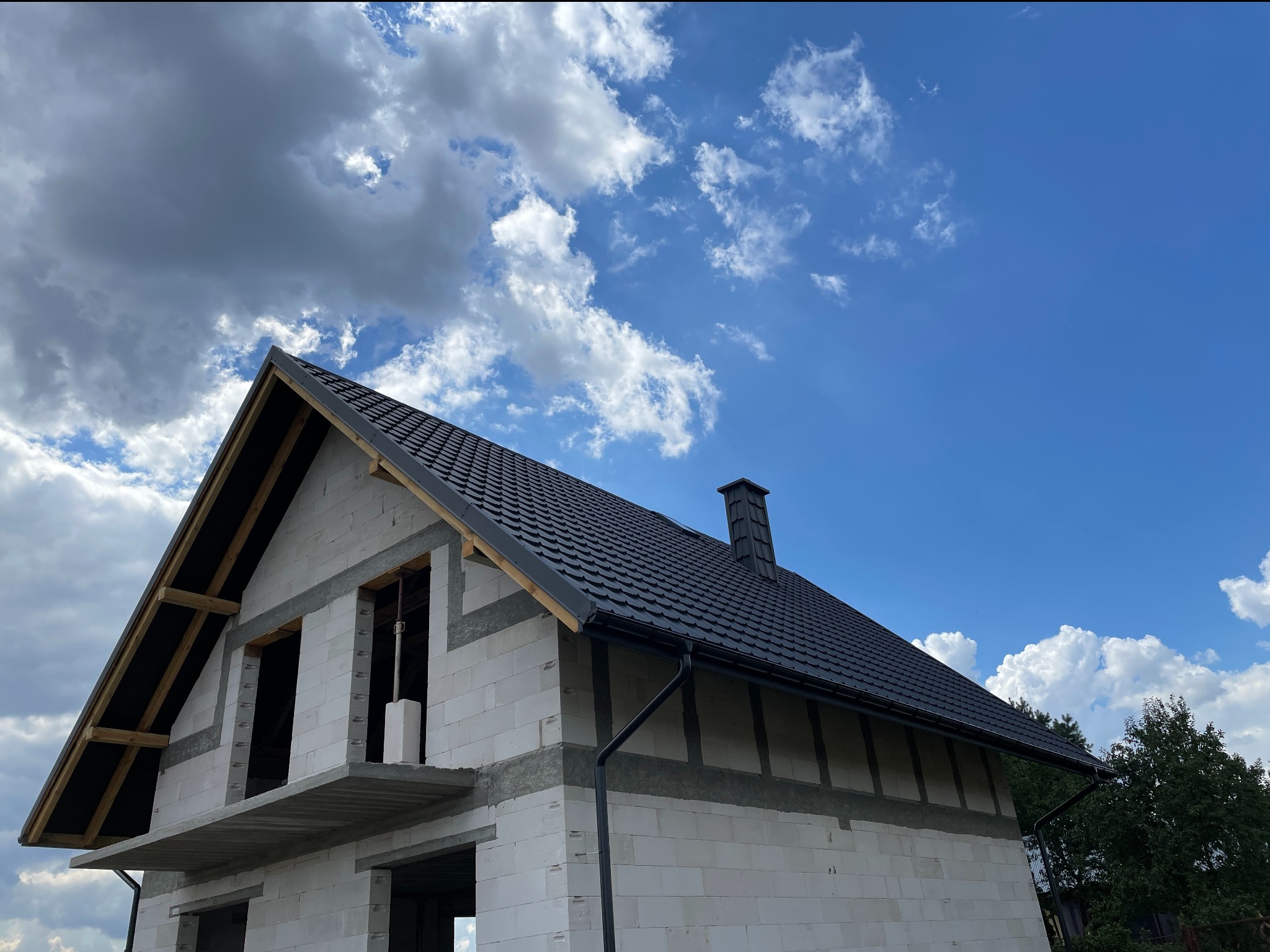 Surowy dom z czarnym dachem w budowie, widok z dołu na tle błękitnego nieba z chmurami. Widoczne orynnowanie i komin.