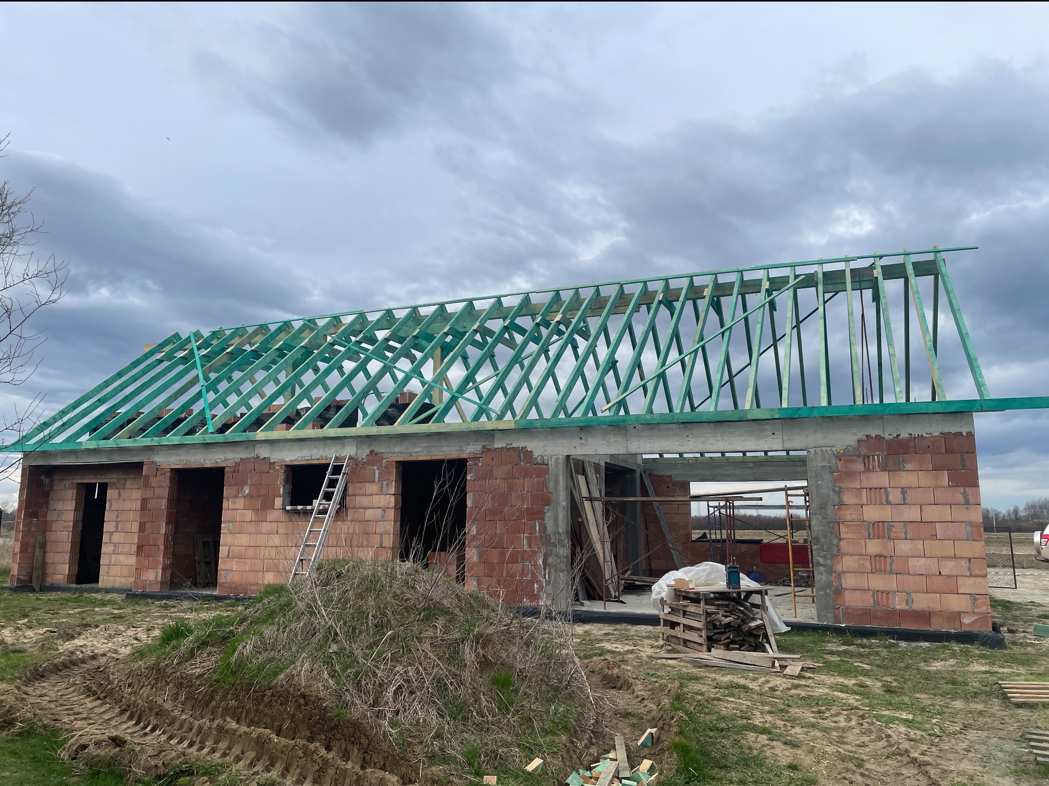 Wznoszona konstrukcja domu z czerwonej cegły i zielonej więźby dachowej pod pochmurnym niebem. Widoczna drabina i materiały budowlane.