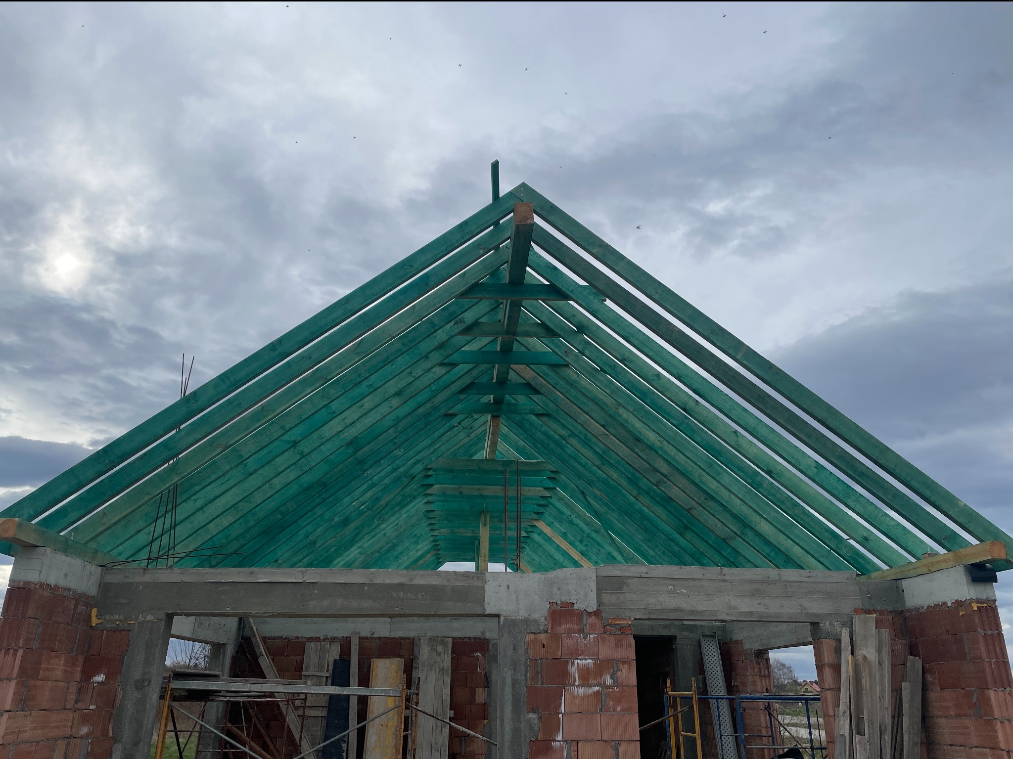 Wznoszenie zielonej więźby dachowej na murowanym budynku w surowym stanie, widok od dołu na tle pochmurnego nieba.