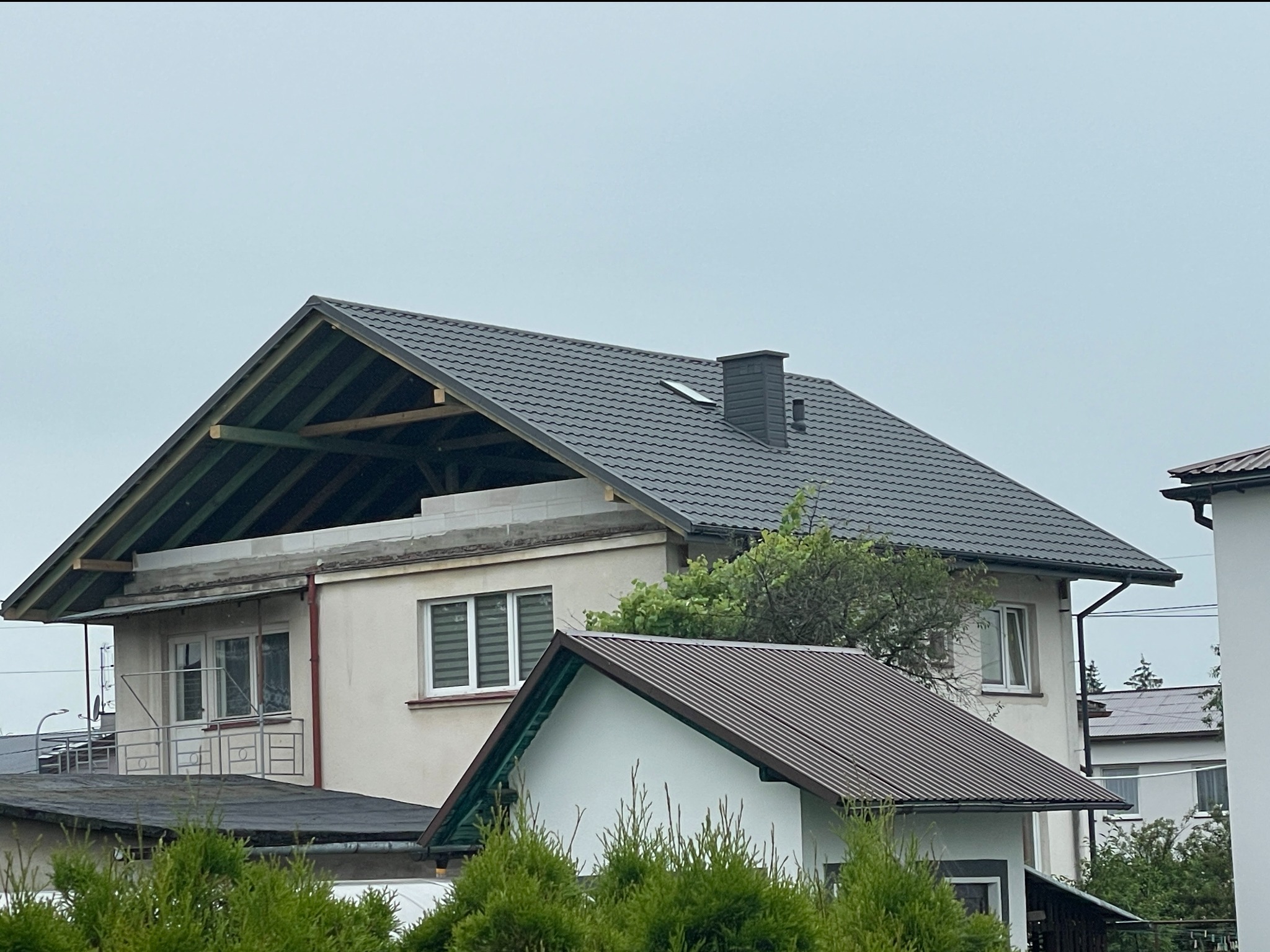 Dom w trakcie prac dekarskich: nowa, grafitowa dachówka ułożona na części dachu, widoczna więźba dachowa i komin. Fragment elewacji domu i zieleń w tle.
