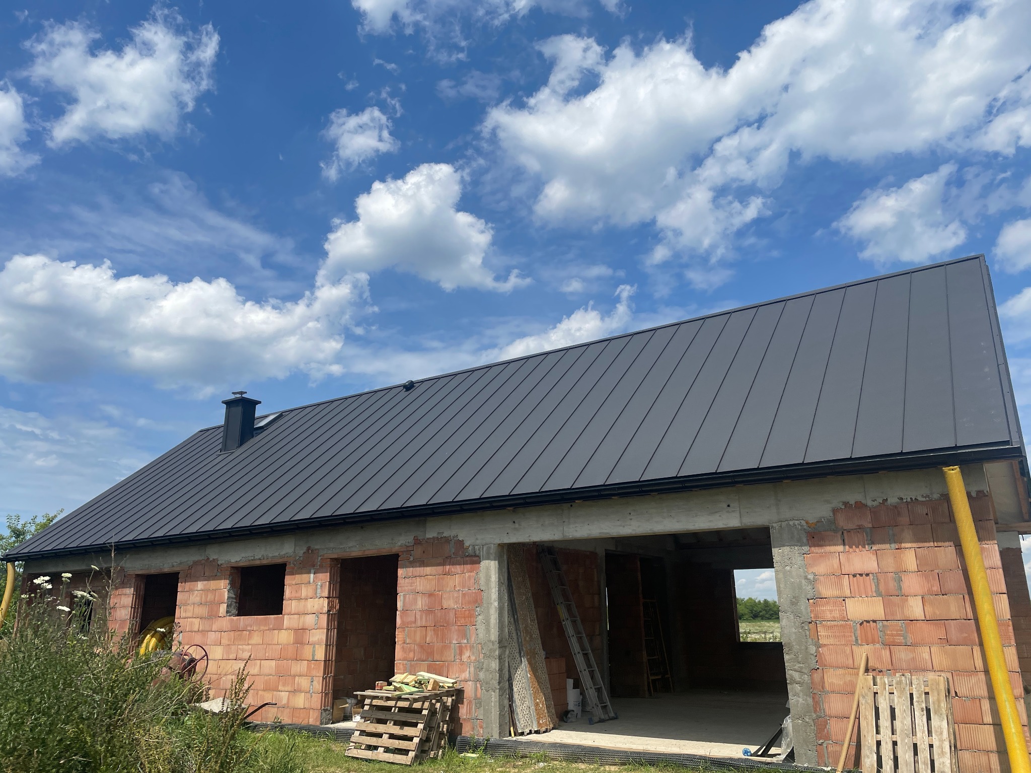 Dom w trakcie budowy z czarnym, metalowym dachem na tle błękitnego nieba z chmurami. Widoczne ceglane ściany i elementy konstrukcyjne.