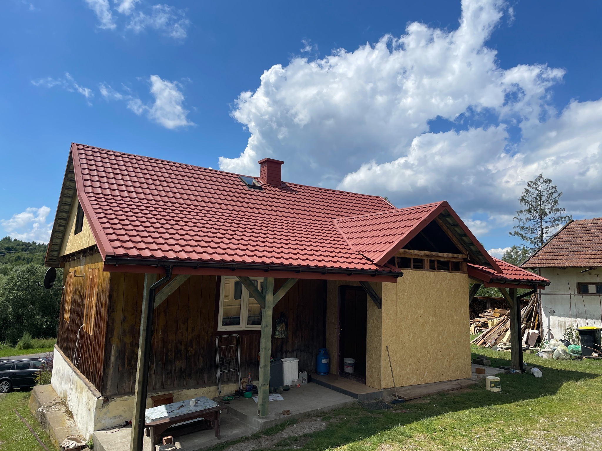 Czerwony dach na drewnianym domu w trakcie prac. Część elewacji obłożona płytą OSB, widoczne materiały budowlane. Słoneczny dzień, błękitne niebo z chmurami.