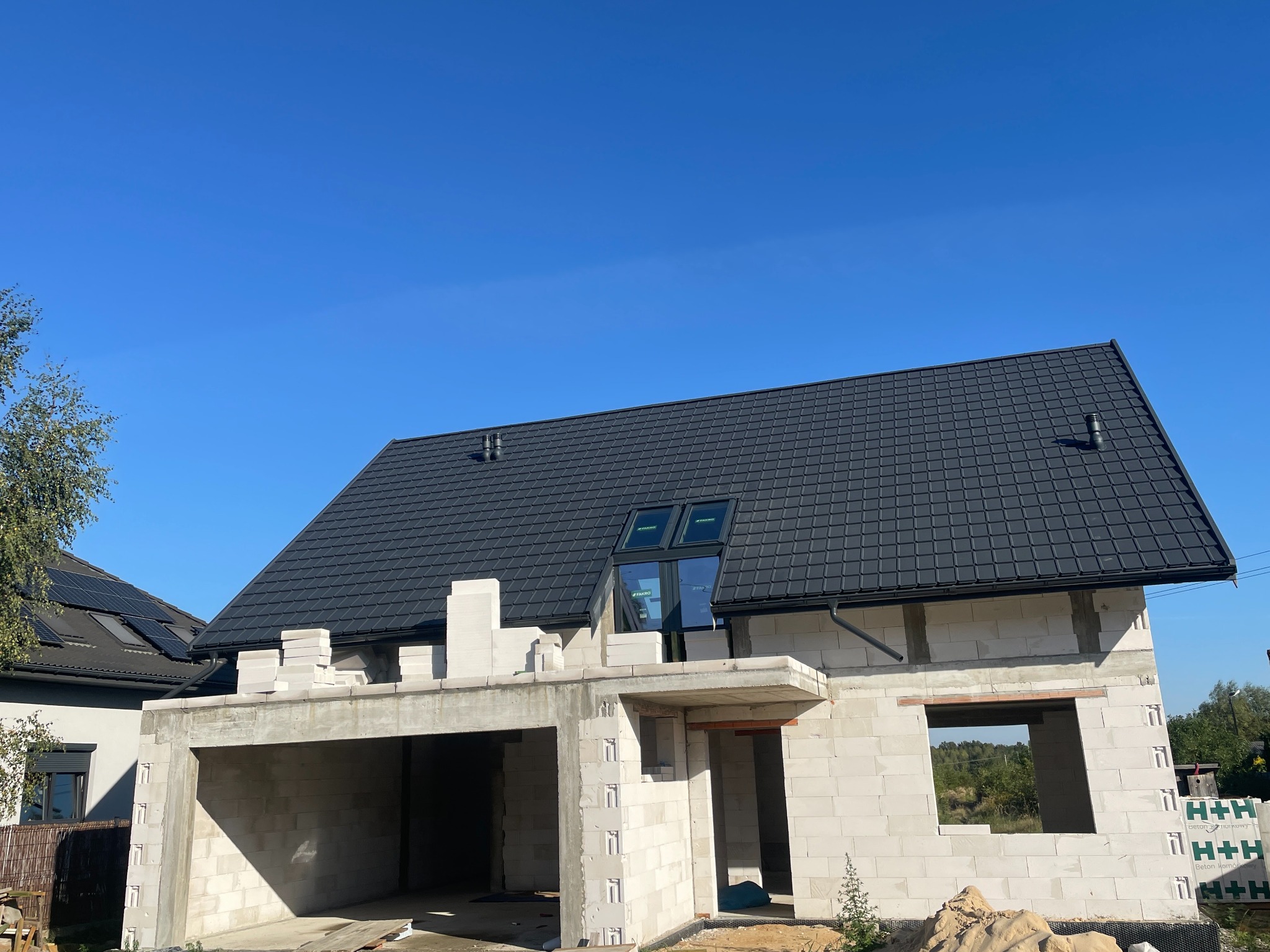 Dom w trakcie budowy z czarnym dachem z dachówki, widoczne okna dachowe i ściany z betonu komórkowego, błękitne niebo w tle.