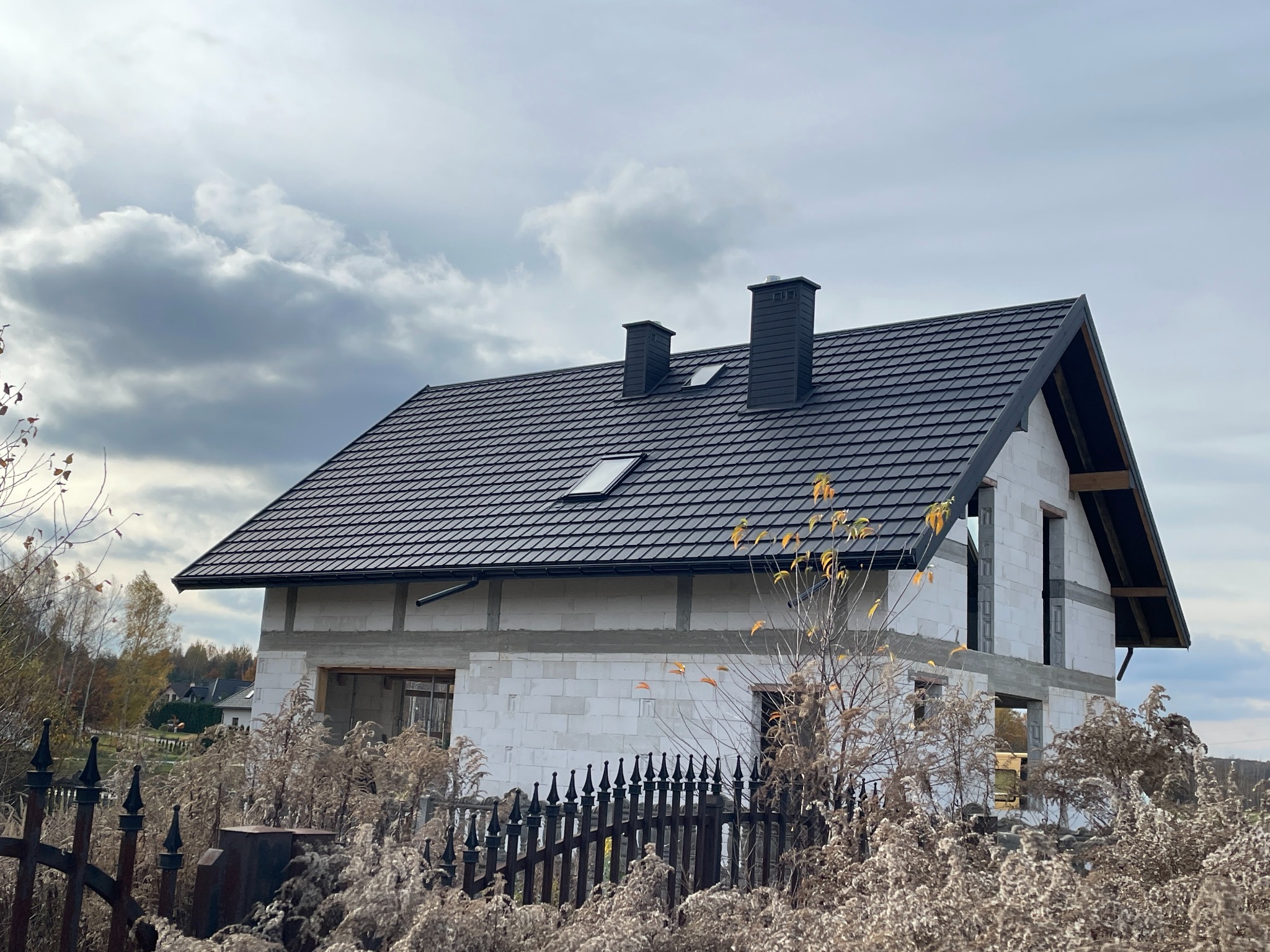 Dom w trakcie budowy z czarnym dachem z dachówki ceramicznej i dwoma kominami, widok zza metalowego ogrodzenia i suchych traw w pochmurny dzień.