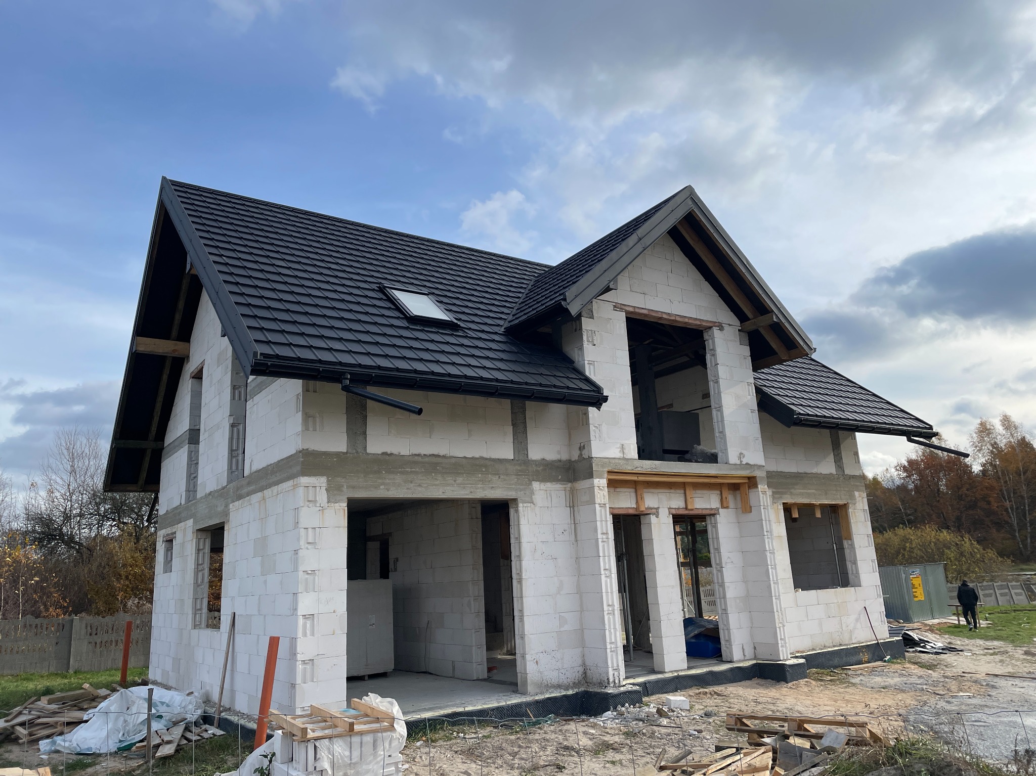 Dom w stanie surowym otwartym z czarnym dachem z dachówki ceramicznej. Widoczne okno dachowe i orynnowanie. Budynek z białego bloczka, w tle drzewa i błękitne niebo.