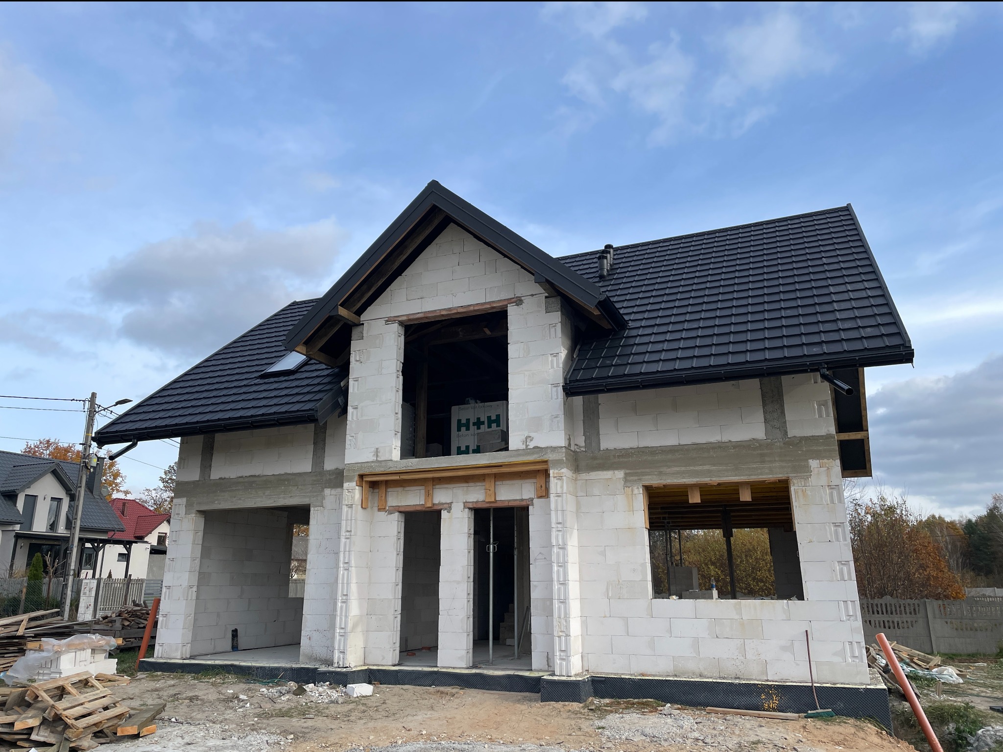 Dom w stanie surowym otwartym z czarnym dachem w Jeżowem. Widoczny pustak biały i drewniane elementy konstrukcji. Dachówka ceramiczna nowa, błyszcząca.