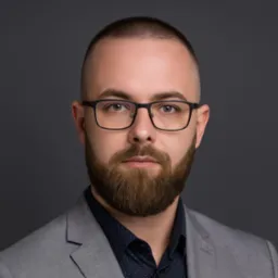 Portret brodatego mężczyzny w okularach i szarej marynarce na ciemnoszarym tle. Profesjonalny wizerunek z wyraźnym spojrzeniem.