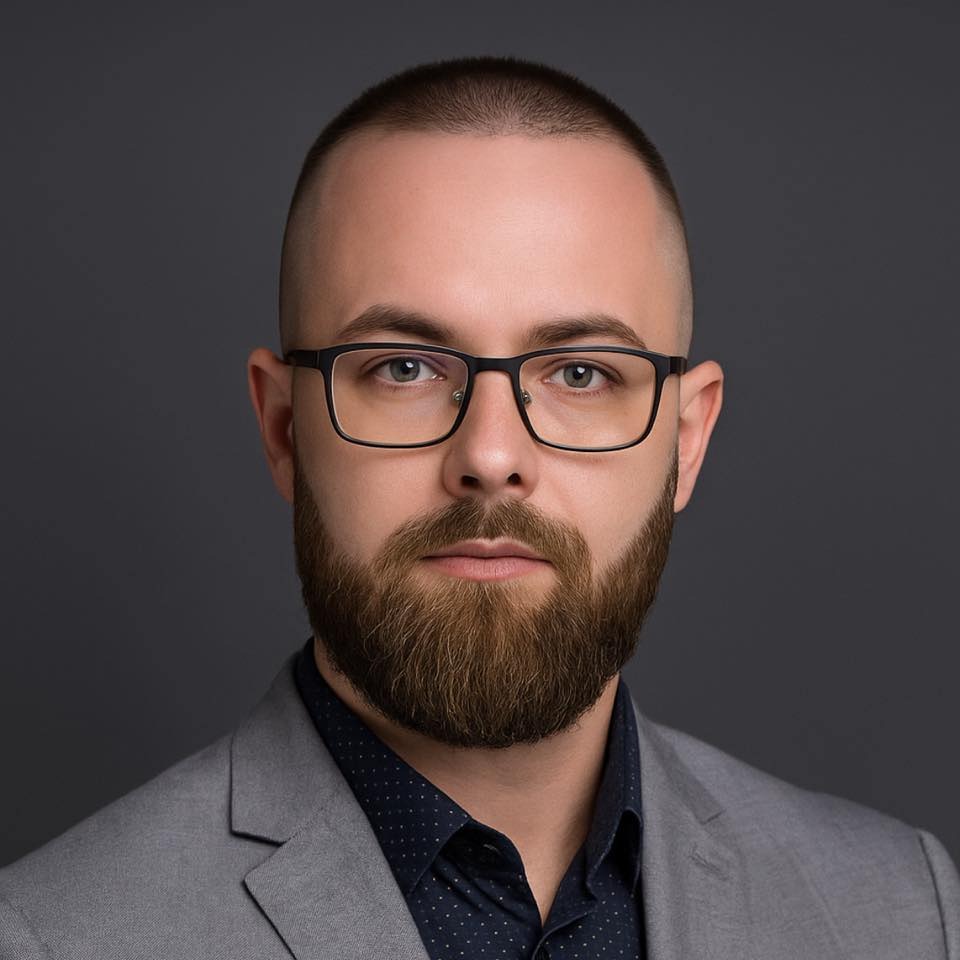 Portret brodatego mężczyzny w okularach i szarej marynarce na ciemnoszarym tle. Profesjonalny wizerunek z wyraźnym spojrzeniem.