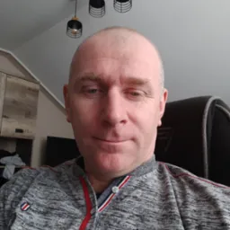 Portret mężczyzny w szarej bluzie, z widocznymi lampami sufitowymi i fragmentem mebli w tle. Ujęcie z bliska, oświetlenie naturalne.