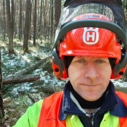 Andrzej Golec - Portret mężczyzny w lesie, w kasku ochronnym Husqvarna i odblaskowej kamizelce. W tle drzewa i resztki śniegu. Ujęcie z bliska, z widocznym wyrazem twarzy.