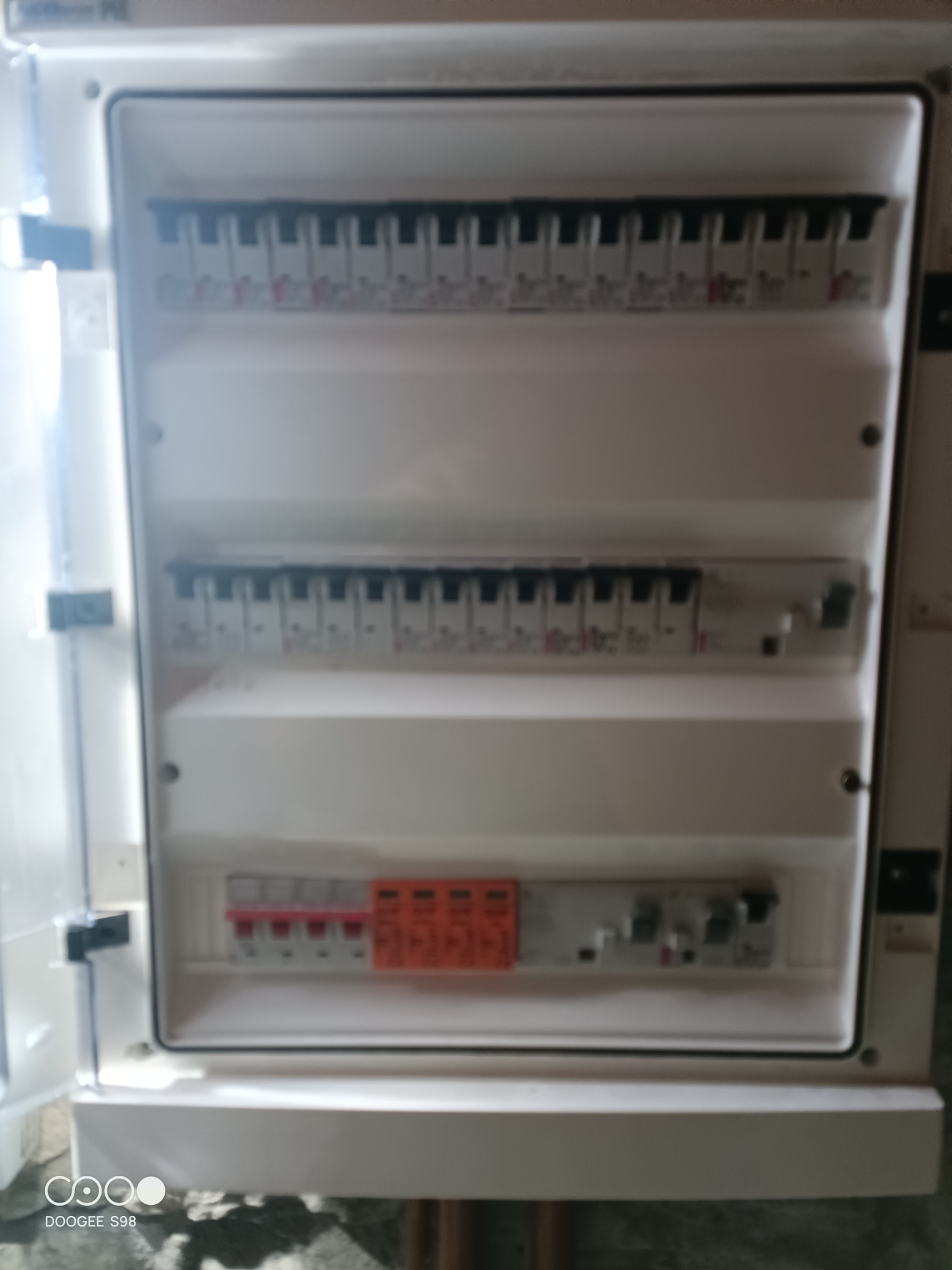 Otwarta skrzynka bezpiecznikowa z widocznymi bezpiecznikami i wyłącznikami, zamontowana na ścianie. Widoczne trzy rzędy elementów.