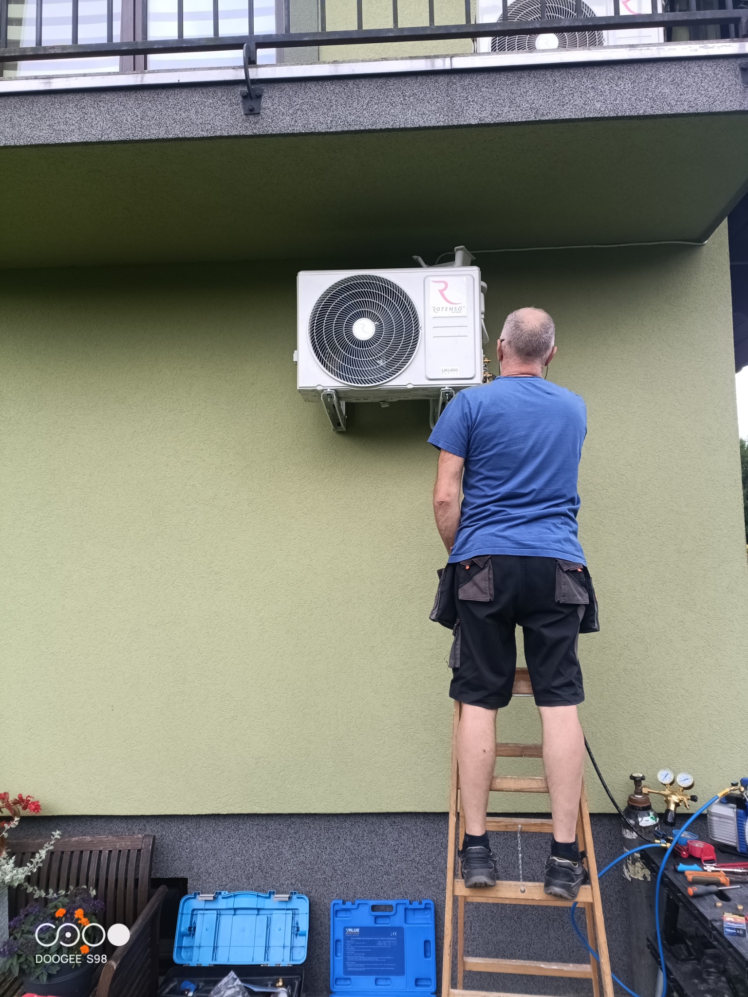 Montera na drabinie montuje klimatyzator Rotenso na elewacji domu. Widoczne narzędzia, butla z gazem. Ściana w odcieniach zieleni, balkon na górze.