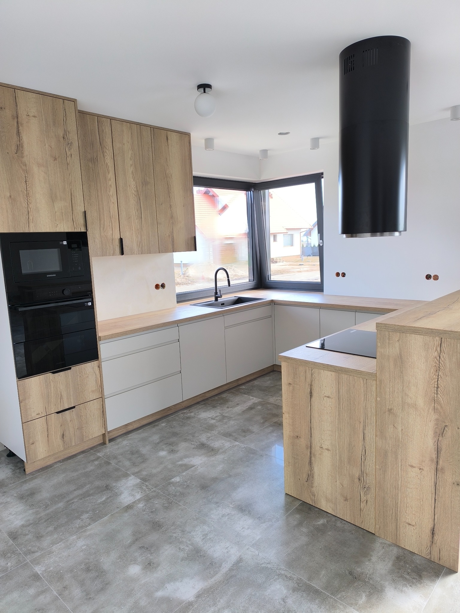 Nowoczesna kuchnia z drewnianymi frontami i szarymi szafkami, czarny okap cylindryczny, płyta indukcyjna i piekarnik w zabudowie. Minimalistyczne oświetlenie.