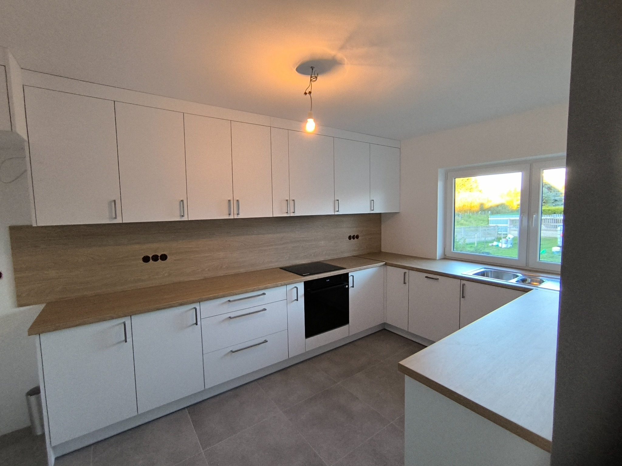 Jasna kuchnia z białymi szafkami i drewnianym blatem, okap z gniazdkami, nowoczesna lampa wisząca. Widok na okno i ogród.